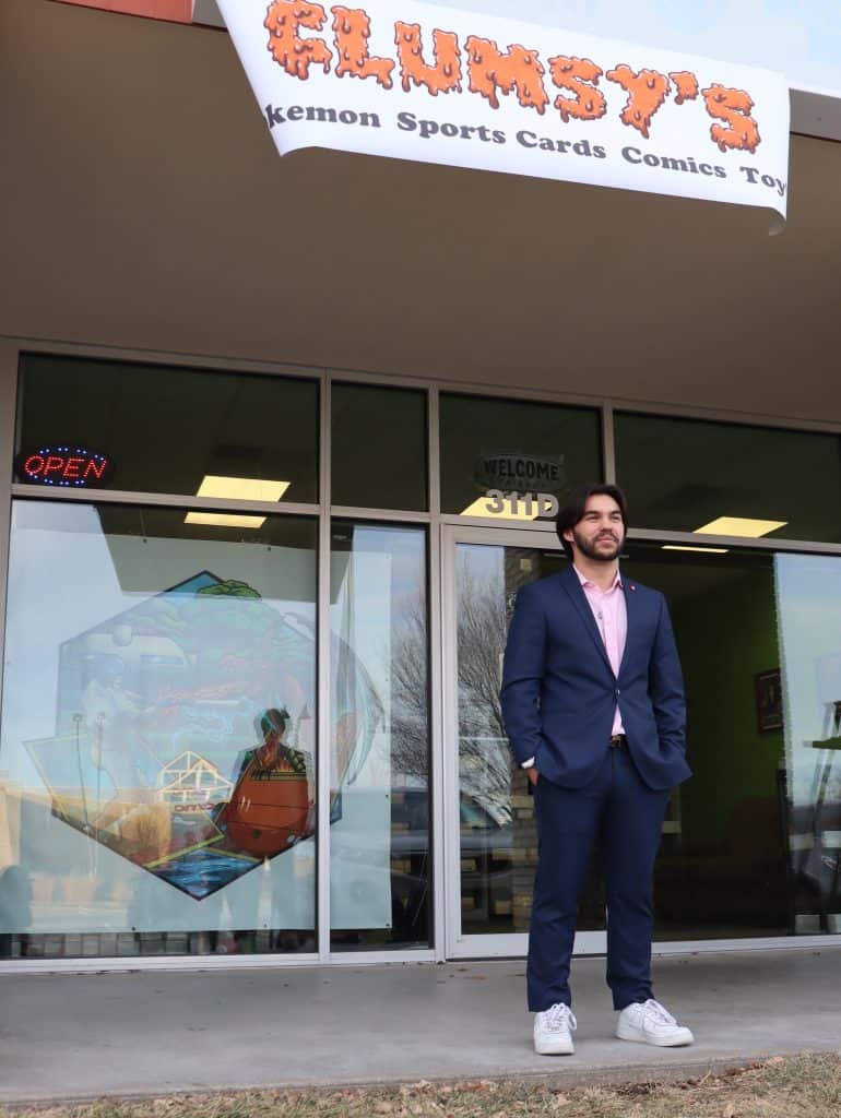 A man in a blue suit and white sneakers stands in front of a storefront with large glass windows on a clear day. Above him, a sign reads "GLUMSY'S [unreadable text] Sports Cards Comics [unreadable text]." A neon "OPEN" sign is visible inside.