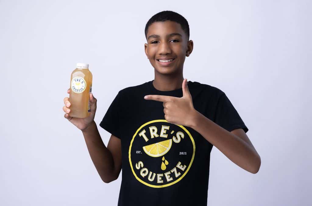 A young person wearing a black "TRE'S SQUEEZE" t-shirt holds a bottle of iced tea in one hand and points at it with the other hand while smiling. The bottle has a label that says "TRE'S SQUEEZE". The background is plain and light-colored.