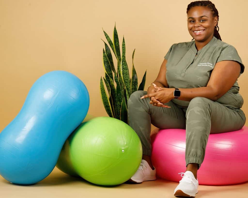 A woman sits on a pink exercise ball, wearing an olive green workout outfit and white sneakers. She is smiling, has braided hair, and a smartwatch on her wrist. Beside her are a blue peanut-shaped exercise ball, a green exercise ball, and a tall houseplant.