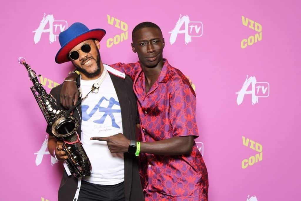 Two men pose in front of a pink backdrop with "A TV" and "VIDCON" written on it repeatedly. The man on the left wears a blue hat with a red band, sunglasses, and a white shirt under a black jacket. He holds a saxophone. The man on the right wears a red and blue patterned outfit.