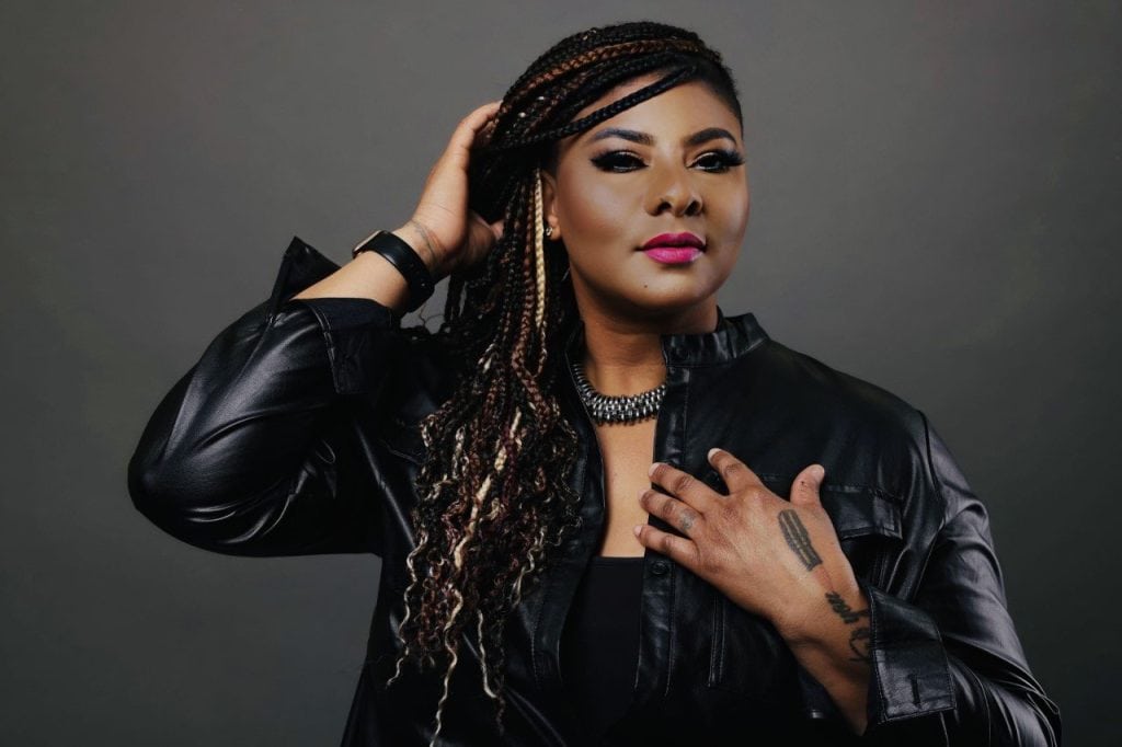 A person with long braided hair poses confidently. They are wearing a black leather jacket, a chunky necklace, and dark eye makeup with bright pink lipstick. One hand is touching their hair, and the other hand is placed on their chest. The background is neutral.
