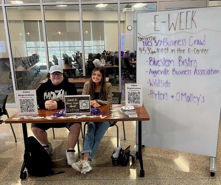 Two people sit at an information table with pamphlets, registration sheets, and QR codes. A whiteboard behind them lists events for "E-Week," including a Business Crawl and visits to local businesses. The setting appears to be an indoor common area.
