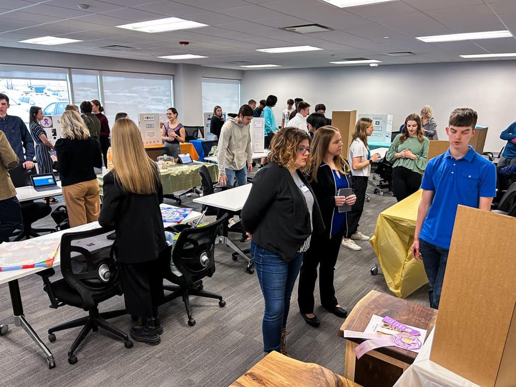 A crowded room with people engaged in discussions and observing presentations. Tables are set up with various displays. Individuals are standing or seated, attentively interacting with exhibits. The room has large windows and a modern office setting.