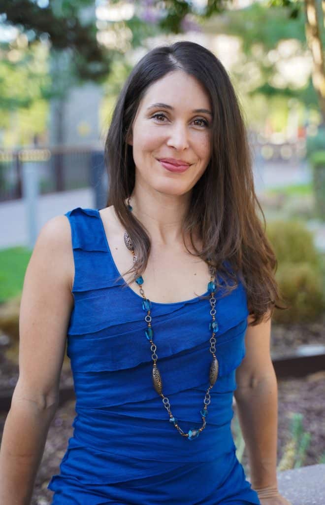 A woman with long brown hair, wearing a blue sleeveless dress and a long necklace, is sitting outdoors. Trees and a blurred urban background are visible. She is smiling and looks relaxed.