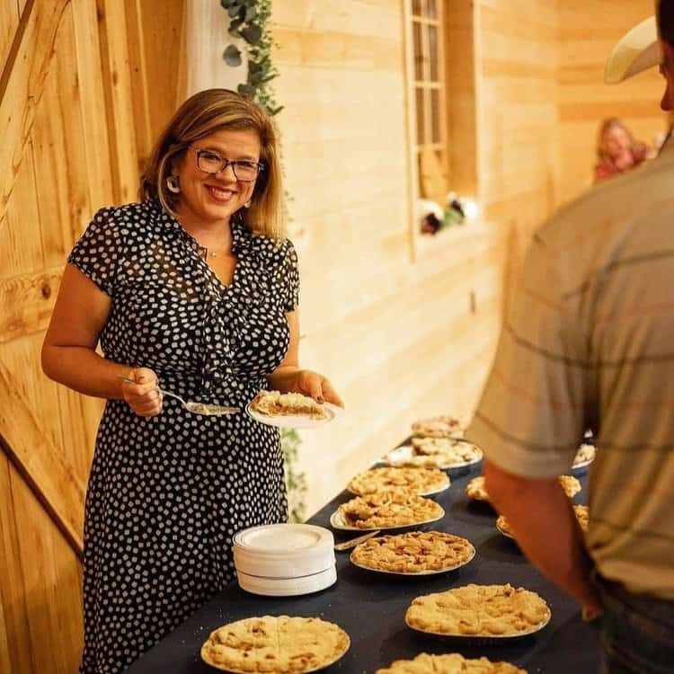 A woman in glasses, wearing a polka-dotted dress, smiles while serving pie at a table that features multiple pies in a warmly lit wooden room. Another person is partly visible in the foreground with their back turned.