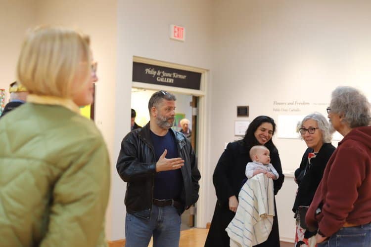 A group of people is gathered in a well-lit gallery space. One man in a black jacket is gesturing with his hand while speaking to others. A woman is holding a baby wrapped in a blanket. Various paintings and a sign indicating a specific gallery wing are visible in the background.
