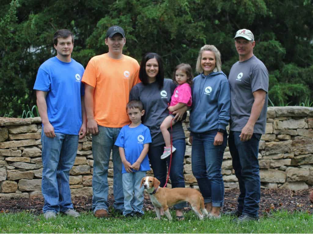 A group of seven people and a dog are posing outdoors in front of a stone wall and greenery. There are three men, two women, and two children. One man wears an orange shirt, another a blue one, and the third a grey one. The children and women wear various clothes. A dog stands in front.