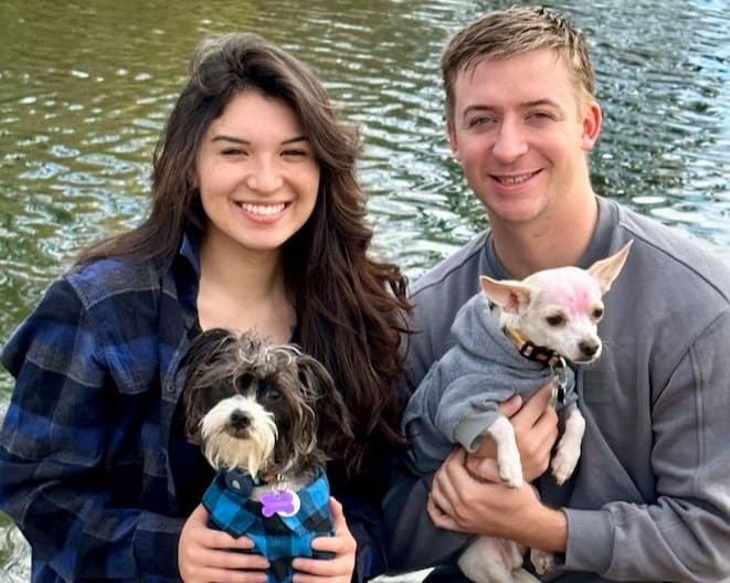 A woman and a man smile while sitting in front of a body of water, each holding a small dog. The woman, in a plaid shirt, holds a small black and white dog wearing a blue shirt. The man, in a gray sweatshirt, holds a small white dog with pink markings wearing a gray shirt.
.