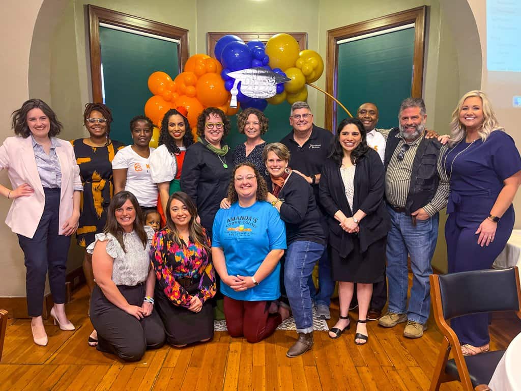 A group of 15 people stands together, smiling at the camera in a room with wooden floors. Behind them is a colorful balloon arch. The individuals are dressed in a mix of casual and business attire. Some are standing while others kneel in the front row.