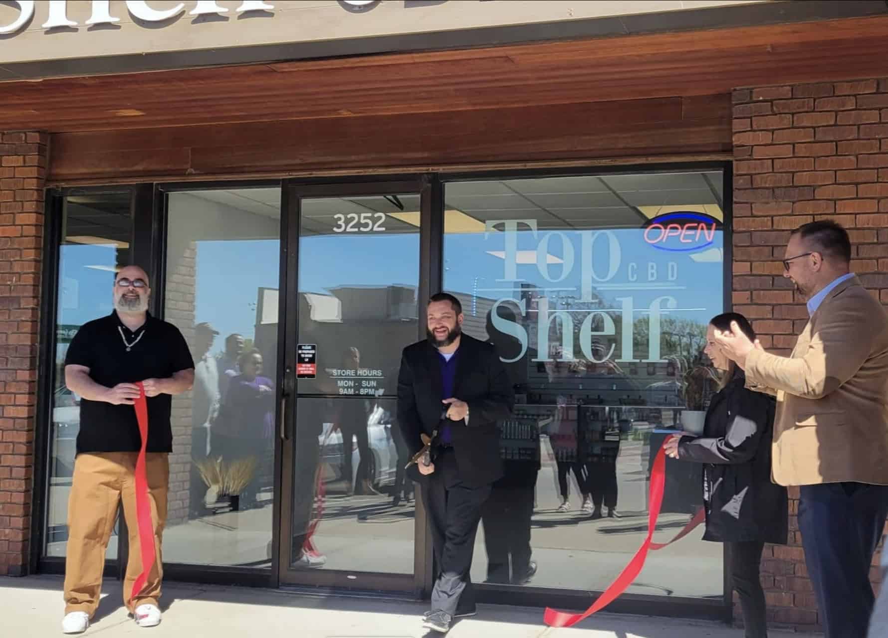 Four people stand in front of a storefront with "Top Shelf" on the window. Two men holding red ribbons stand on either side of a man in the center holding scissors, about to cut the ribbon. Another man and a child standing on the right applaud. The store sign reads "3252".