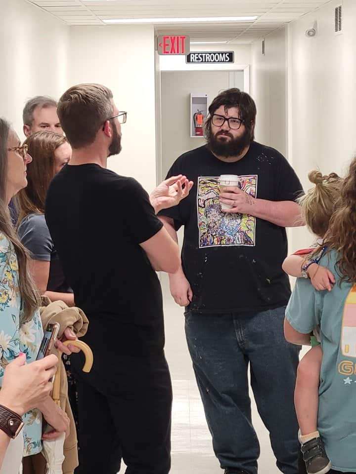 A group of people is standing in a hallway under fluorescent lighting, conversing. One person with a beard and glasses is holding a cup and wearing a black shirt, while others listen attentively. In the background, a restroom sign is visible above a door.
