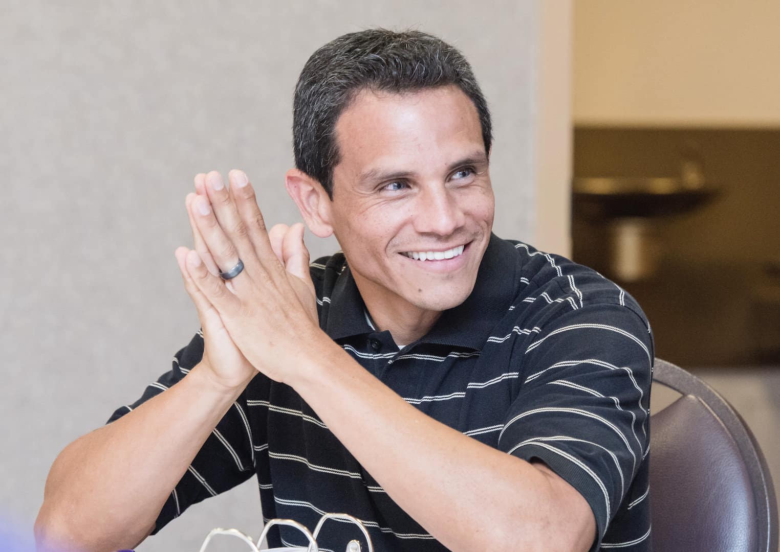 A man with short dark hair and wearing a black striped polo shirt is sitting and smiling, his hands clasped together in front of him. The background is slightly blurred, featuring neutral-colored walls and a glimpse of a darker corridor.