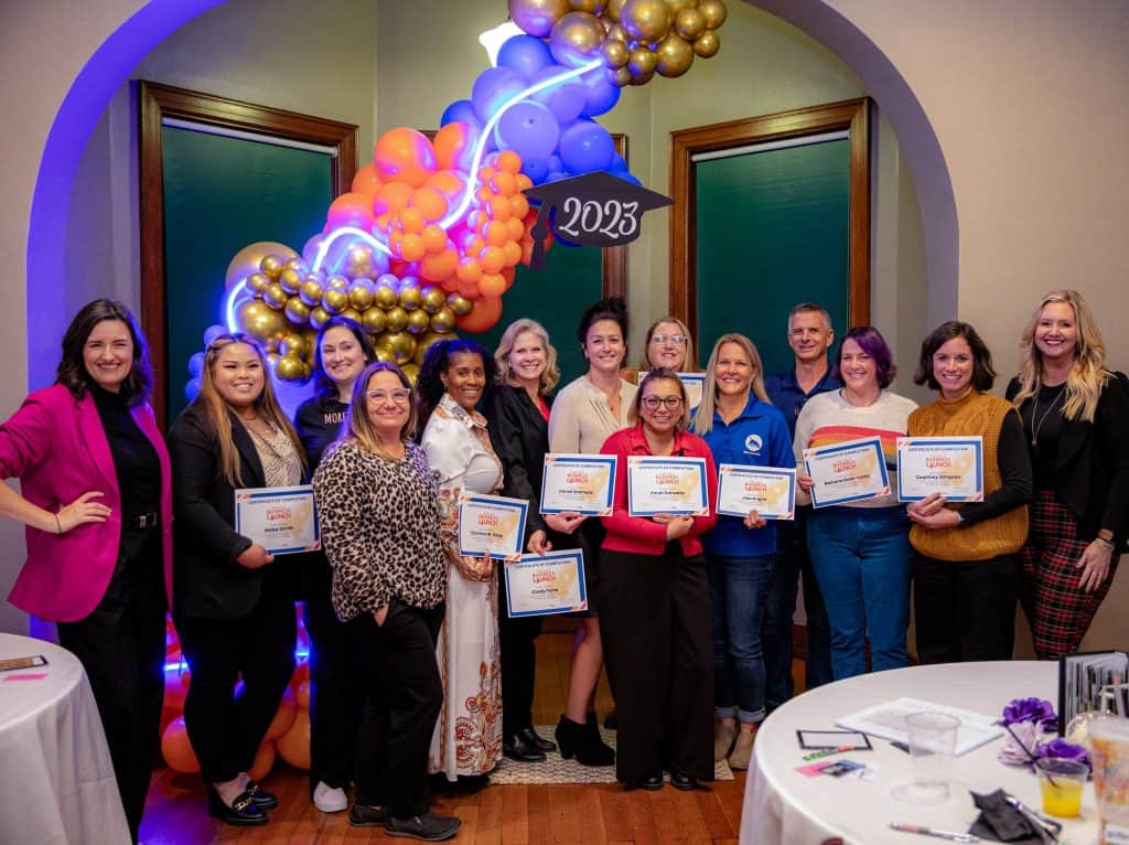 A group of people pose for a photo at an event in front of a balloon arch with the year "2023" displayed. They are holding certificates and smiling. The room is decorated in bright colors, and a table with papers and glasses is partially visible in the foreground.