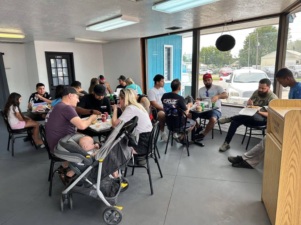 A diverse group of people sits at tables inside a casual eatery. Some are eating food, others are chatting. A baby in a stroller is in the foreground. Large windows show the street and parked cars outside. The atmosphere appears friendly and lively.
