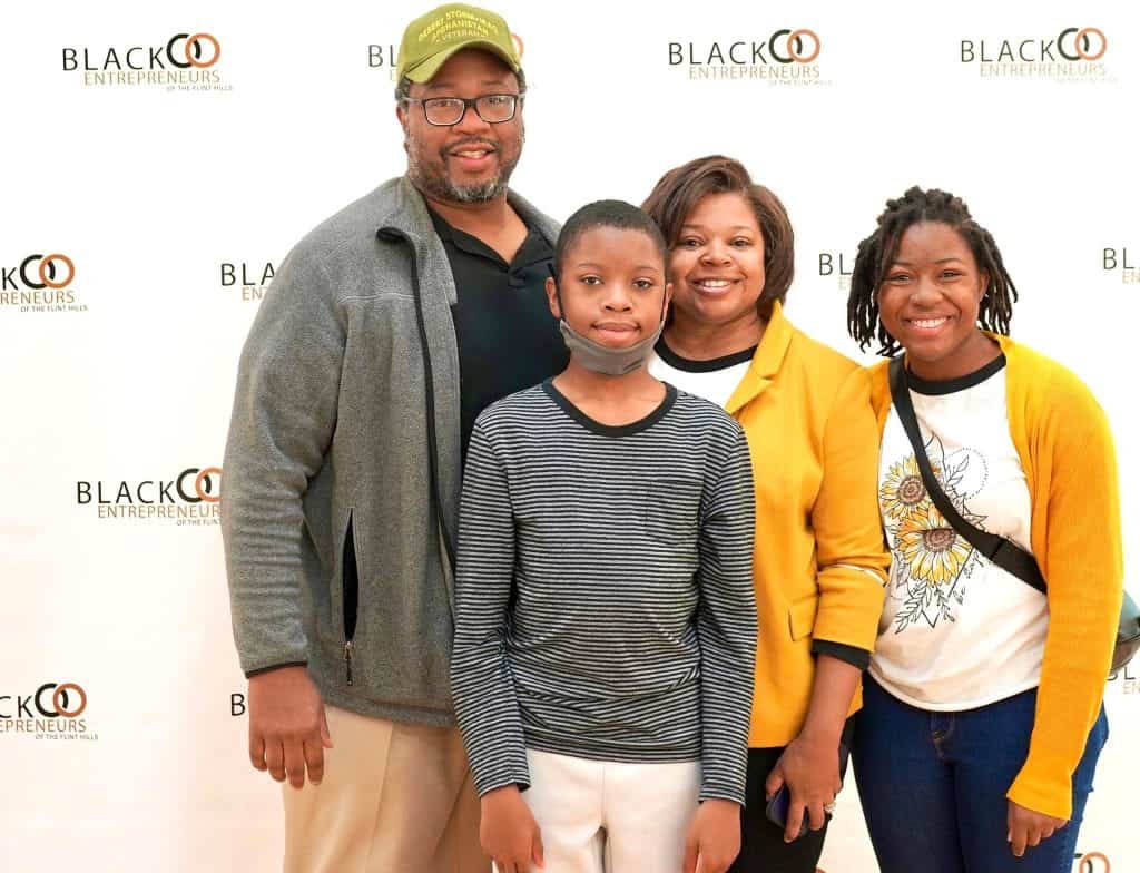 A group of four people posing for a photo in front of a background with the logo "Black Entrepreneurs". The group includes an adult man in a green cap, an adult woman in a yellow cardigan, a younger boy in a striped shirt, and a young woman in a white shirt.