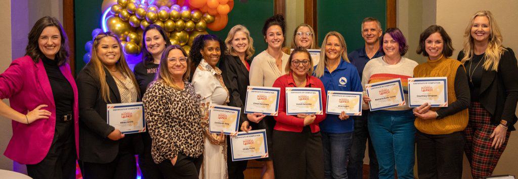 A group of 14 people stands together, smiling and posing for a photo. Several individuals in the group are holding certificates. The background features balloons and decorations, suggesting a celebratory event.