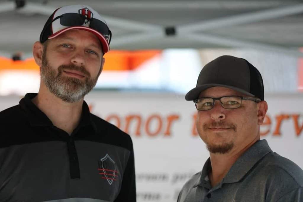 Two men stand side by side under a canopy. Both are wearing hats and glasses. The man on the left has a beard and wears a black shirt with a logo. The man on the right has a goatee and wears a gray shirt. A blurred sign in the background partially reads "honor".