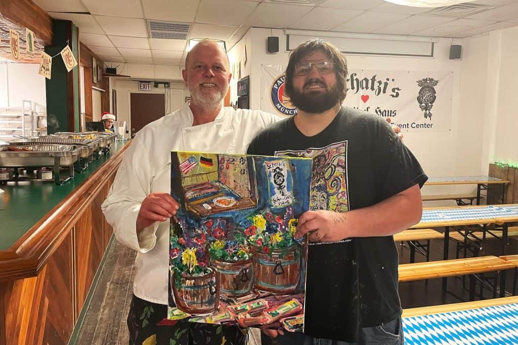 Two men stand in a restaurant, smiling at the camera. One, in a chef's coat, stands on the left with his arm around the other man, who holds a colorful painting of flowers and barrels. The restaurant features wooden counters and white walls with posters.