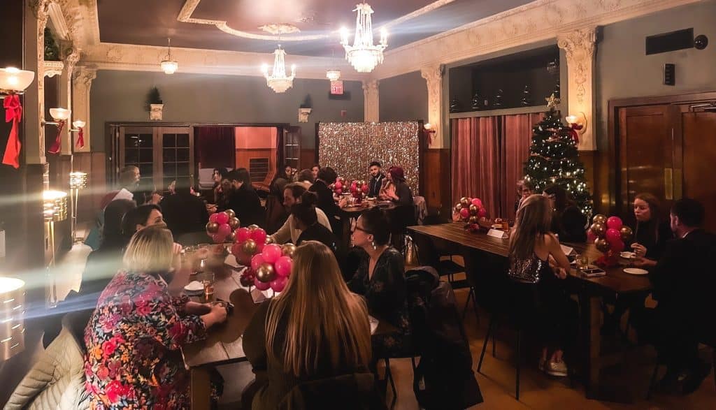 A festive gathering takes place in a warmly lit room with ornate ceiling decorations. Tables are adorned with red and gold balloon centerpieces. A decorated Christmas tree stands in the corner. Attendees are engaged in conversations, enjoying the holiday ambiance.