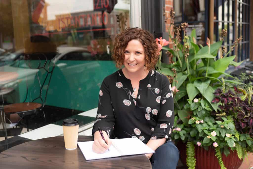 A person with curly hair, wearing a black blouse with floral patterns, sits at an outdoor table of a café. They are smiling and writing in a notebook. A cup of coffee is placed on the table beside them, and there are green plants in the background.