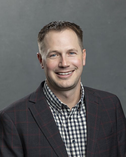 Brian Rempe, a man with short brown hair, smiles against a gray background. He is wearing a black and red checkered blazer over a black and white checkered shirt.