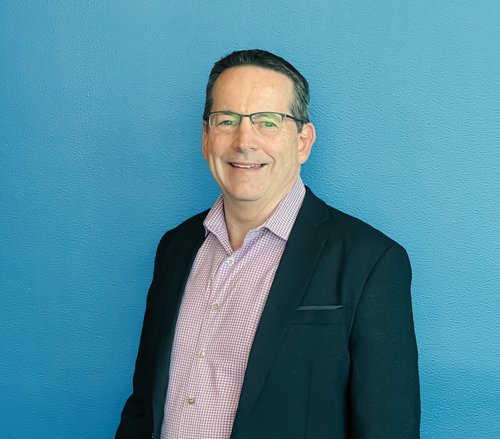 A man with short, dark hair and glasses is smiling while wearing a black blazer over a pink checkered shirt. He looks like he just finished an Auto Draft as he stands against a solid blue background.