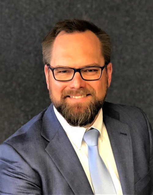 Jeff Wick, a man with a beard and glasses, is smiling at the camera. He is wearing a blue suit, a white shirt, and a blue tie. The background is dark with a textured appearance.
