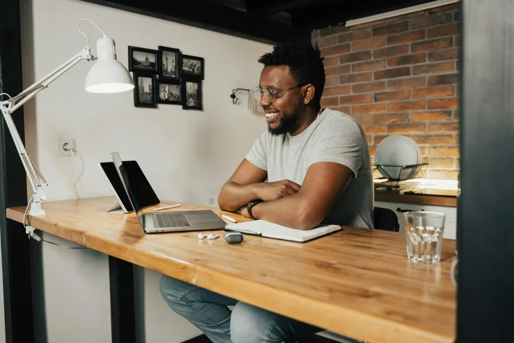 A man sits at a wooden desk with a laptop, tablet, and notebooks. He is wearing a gray t-shirt and glasses, smiling while looking at the laptop. The desk has a lamp, a glass of water, and a mouse. The background features a brick wall and framed photos.