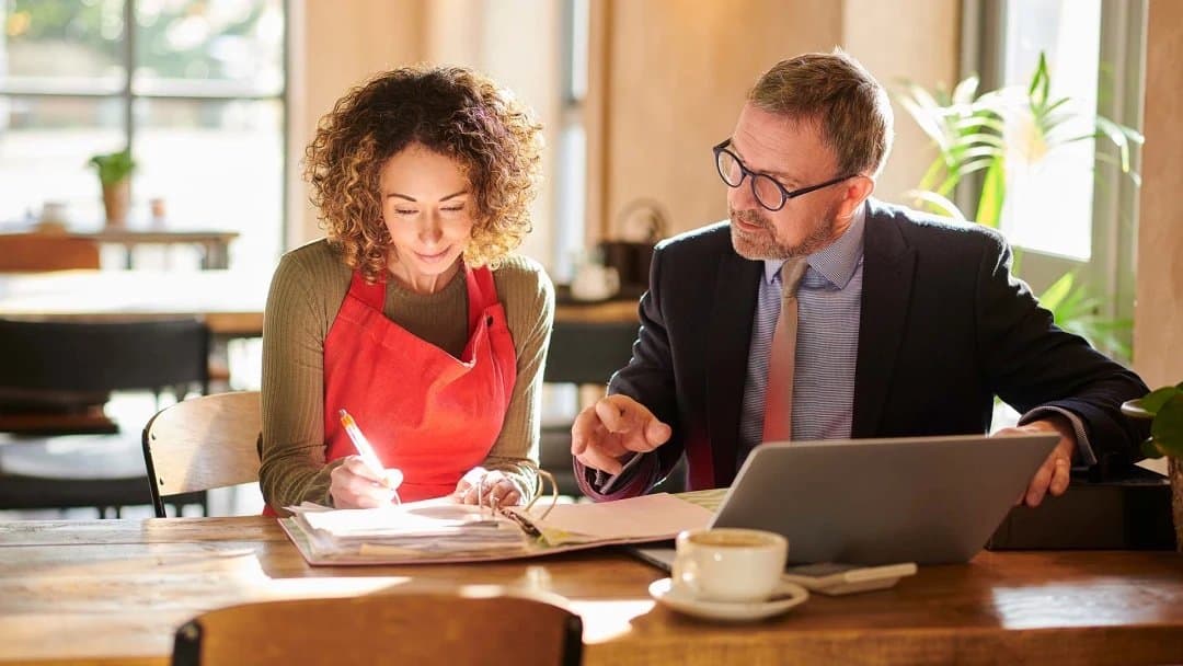 A woman in a red apron and a man in a suit are sitting at a wooden table in a bright, sunlit room. The woman is writing in a notebook while the man, with a laptop open, points to the notebook. A coffee cup is on the table in front of them.