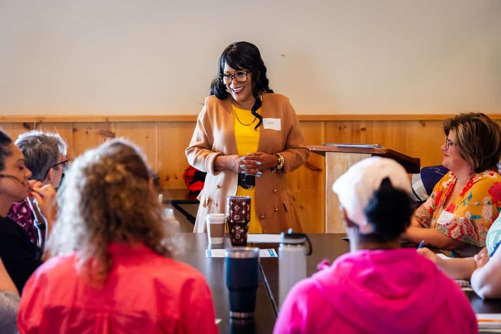 A woman in a beige blazer and yellow top stands at the front of a room, smiling, and speaking to a group of seated people. The attendees are listening attentively around a table, engaged in the presentation. A lectern is visible beside the speaker.