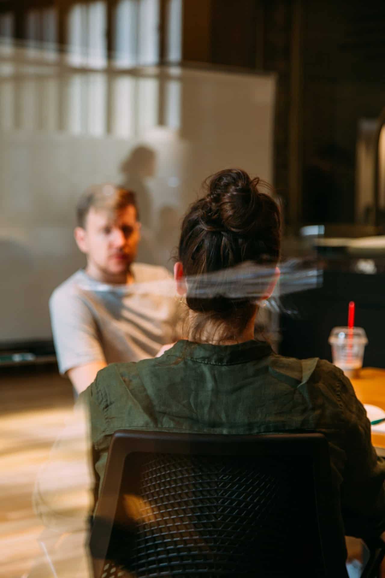 A woman with a bun sits across a table from a man with a beard, engaged in conversation. They are in a well-lit room with a large window and a reflective surface, possibly a glass wall, behind them. There is a drink with a red straw on the table.
