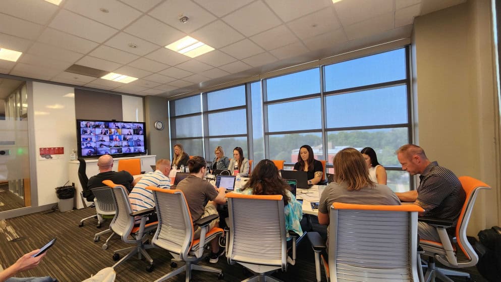 A diverse group of people is seated around a large conference table in a modern office with large windows. They are engaged in a meeting, some using laptops. A screen on the wall shows a video conference with participants in small windows.